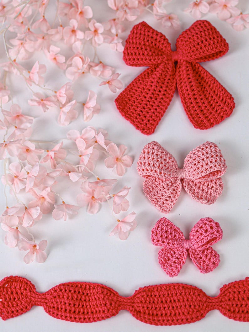 May include: Three crochet bows in varying shades of red and pink, arranged vertically. The top bow is a deep red, the middle is a soft light pink, and the bottom is a vibrant pink. A long, scalloped red crochet trim piece lies horizontally below.