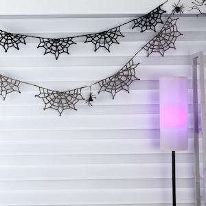 May include: Two rows of crocheted spider web bunting hanging horizontally. The top row is black, and the bottom row is a light gray. Small black plastic spider figures are attached to the web patterns at various intervals along the string.