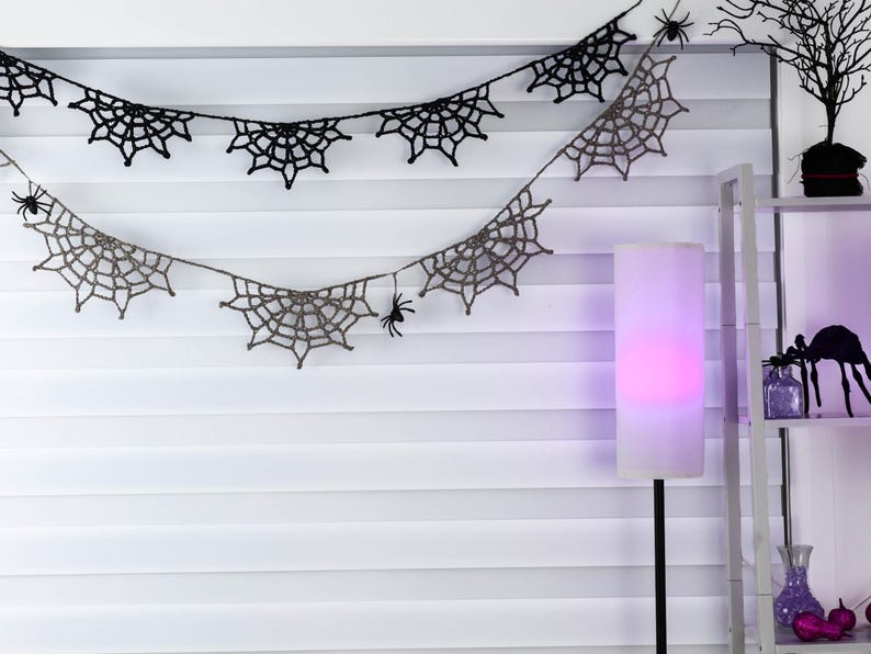 May include: Two rows of crocheted spider web bunting hanging horizontally. The top row is black, and the bottom row is a light gray. Small black plastic spider figures are attached to the web patterns at various intervals along the string.