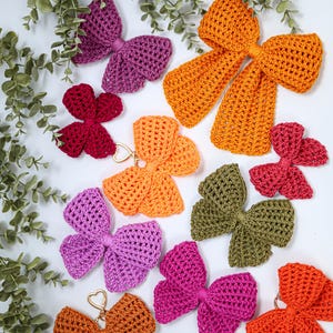 May include: A collection of crocheted bows in various colors including orange, purple, magenta, olive green, and dark red. Some bows feature gold-toned heart-shaped metal clasps attached to the center, while others are plain fabric bows.