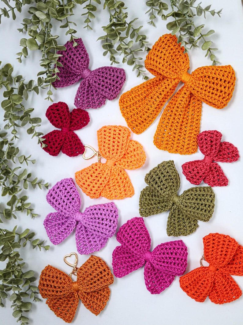 May include: A collection of crocheted bows in various colors including orange, purple, magenta, olive green, and dark red. Some bows feature gold-toned heart-shaped metal clasps attached to the center, while others are plain fabric bows.