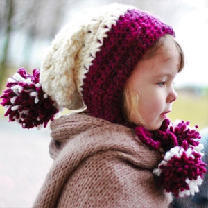 May include: A young girl wearing a white and burgundy crocheted hat with a pom-pom on top. The hat has a long, crocheted scarf attached to it. The girl is wearing a brown knitted scarf.