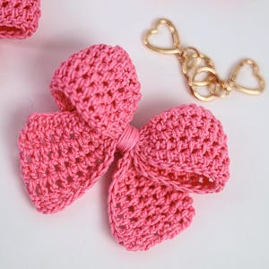 May include: A bright pink crocheted bow accessory featuring a textured yarn construction with a central knotted detail. Beside the bow lies a gold-toned metal keychain hardware piece that includes two heart-shaped clasps and circular rings.