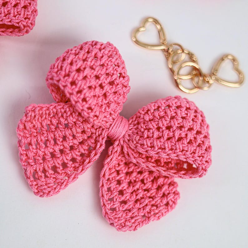 May include: A bright pink crocheted bow accessory featuring a textured yarn construction with a central knotted detail. Beside the bow lies a gold-toned metal keychain hardware piece that includes two heart-shaped clasps and circular rings.