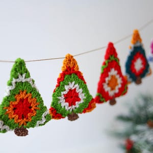 Puede incluir: Una guirnalda de &aacute;rboles de Navidad de ganchillo en verde, rojo, naranja, blanco y azul. Los &aacute;rboles est&aacute;n unidos con un cordel y cuelgan de una pared blanca.
