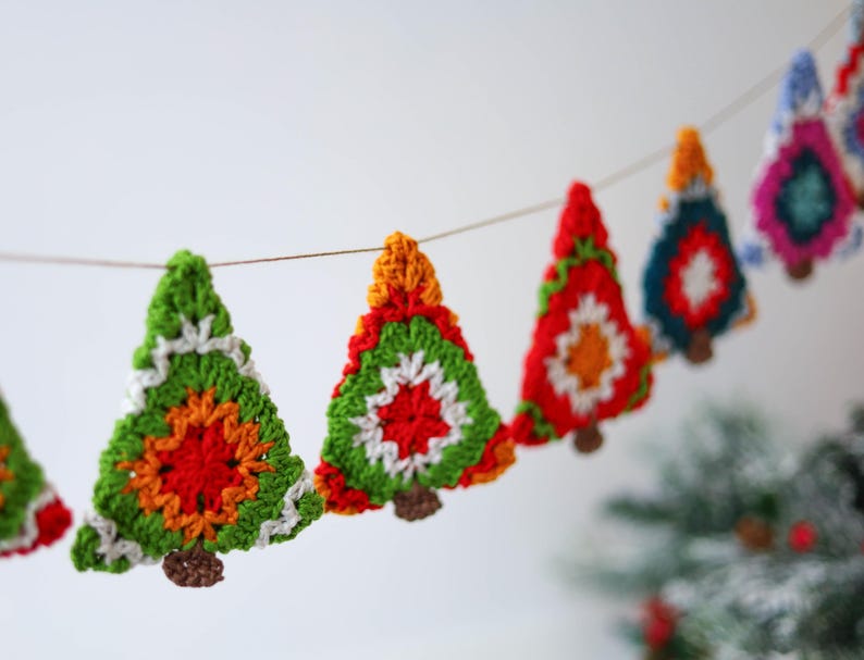 Puede incluir: Una guirnalda de &aacute;rboles de Navidad de ganchillo en verde, rojo, naranja, blanco y azul. Los &aacute;rboles est&aacute;n unidos con un cordel y cuelgan de una pared blanca.