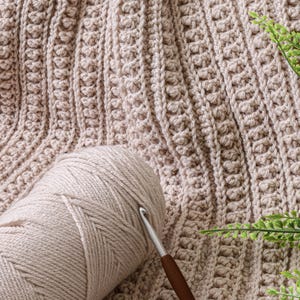May include: A close-up of a beige crochet blanket with a ball of beige yarn and a crochet hook. The blanket has a textured stitch pattern.