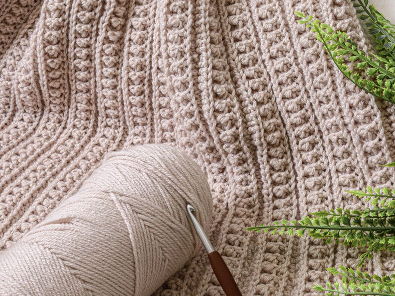 May include: A close-up of a beige crochet blanket with a ball of beige yarn and a crochet hook. The blanket has a textured stitch pattern.