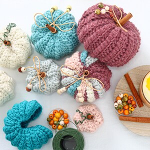 May include: A collection of crocheted pumpkins in various colors including white, teal, light blue, pink, and mauve. Each pumpkin features a cinnamon stick stem, wire tendrils, and wooden beads. A candle, loose beads, and green wire are also present.
