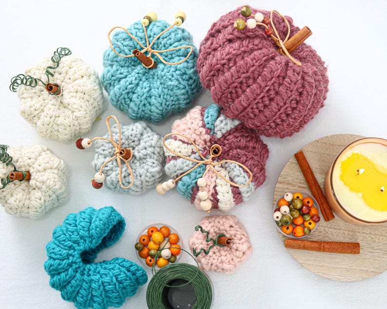 May include: A collection of crocheted pumpkins in various colors including white, teal, light blue, pink, and mauve. Each pumpkin features a cinnamon stick stem, wire tendrils, and wooden beads. A candle, loose beads, and green wire are also present.