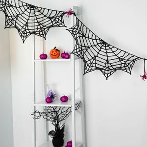 May include: A black crocheted spider web garland featuring small pink plastic spiders attached to the web. The garland is draped across the top of a white shelving unit decorated with small orange and purple pumpkins and a black tree.