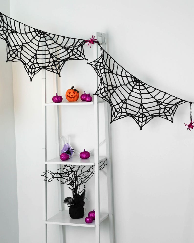 May include: A black crocheted spider web garland featuring small pink plastic spiders attached to the web. The garland is draped across the top of a white shelving unit decorated with small orange and purple pumpkins and a black tree.