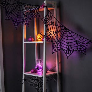 May include: A white wooden ladder shelf decorated with black crocheted spider webs, a glowing orange jack-o'-lantern, purple glitter pumpkins, a clear glass vase with purple lights, and a black decorative tree branch.