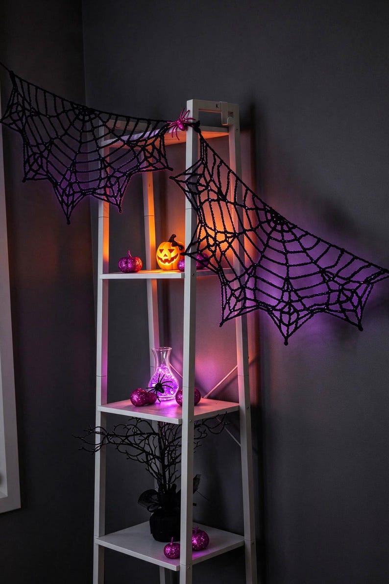 May include: A white wooden ladder shelf decorated with black crocheted spider webs, a glowing orange jack-o'-lantern, purple glitter pumpkins, a clear glass vase with purple lights, and a black decorative tree branch.