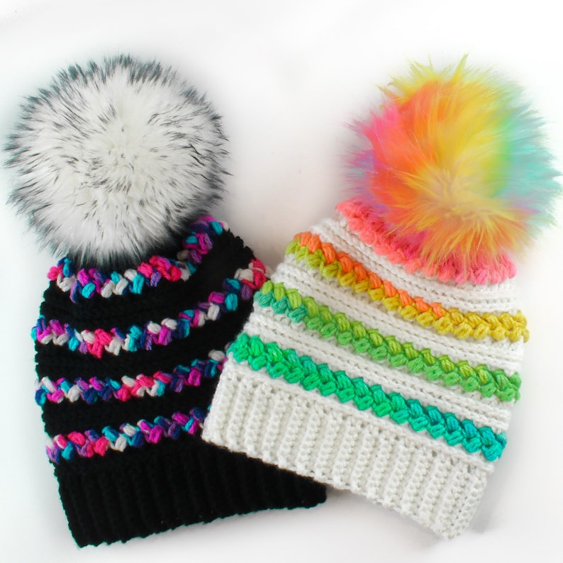 Puede incluir: Dos gorros de ganchillo con pompones de piel sint&eacute;tica. Un gorro es negro con rayas rosas, azules y blancas. El otro gorro es blanco con rayas verdes, amarillas y naranjas.