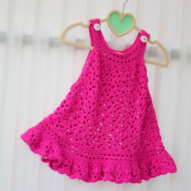 May include: A pink crocheted baby dress with a ruffled hem and white buttons. The dress is hanging on a wooden hanger with a heart-shaped top.