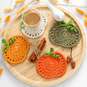 May include: Three crocheted pumpkin-shaped coasters in orange, olive green, and cream colors. The coasters feature a textured stitch pattern with a small green stem. They are arranged on a round wooden tray with cinnamon sticks and acorns.