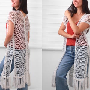 May include: A long, white, open-front crochet cardigan featuring a loose, net-like stitch pattern, short sleeves, and long fringe detailing along the bottom hem. The garment is layered over a solid rust-colored top and light blue denim jeans.
