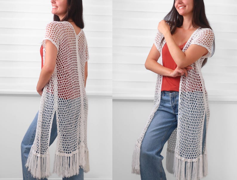 May include: A long, white, open-front crochet cardigan featuring a loose, net-like stitch pattern, short sleeves, and long fringe detailing along the bottom hem. The garment is layered over a solid rust-colored top and light blue denim jeans.