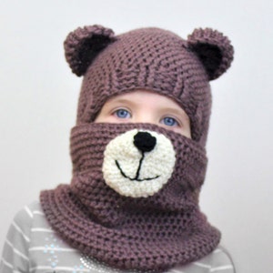 May include: A purple knitted bear hat with a white face and black nose and mouth. The hat has ear flaps and covers the face.