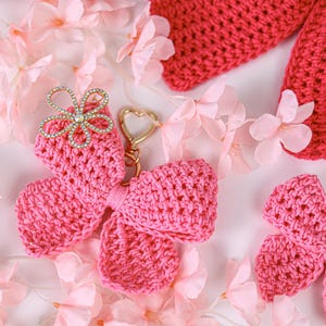 May include: Two bright pink crocheted bow keychains with gold-toned heart-shaped clasps. One bow features a decorative silver-toned flower embellishment with clear rhinestones at the center. The texture is a dense, uniform yarn stitch pattern.