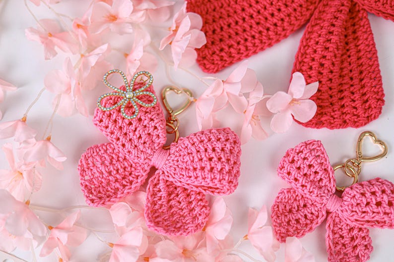 May include: Two bright pink crocheted bow keychains with gold-toned heart-shaped clasps. One bow features a decorative silver-toned flower embellishment with clear rhinestones at the center. The texture is a dense, uniform yarn stitch pattern.