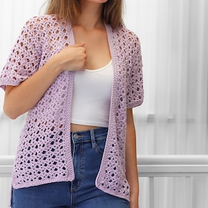 May include: A light purple crochet cardigan with a delicate openwork pattern. The cardigan has a relaxed fit and short sleeves.