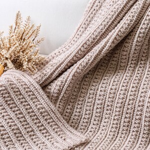 May include: A soft, beige crocheted throw blanket with a textured stitch pattern. The blanket is draped over a white couch.