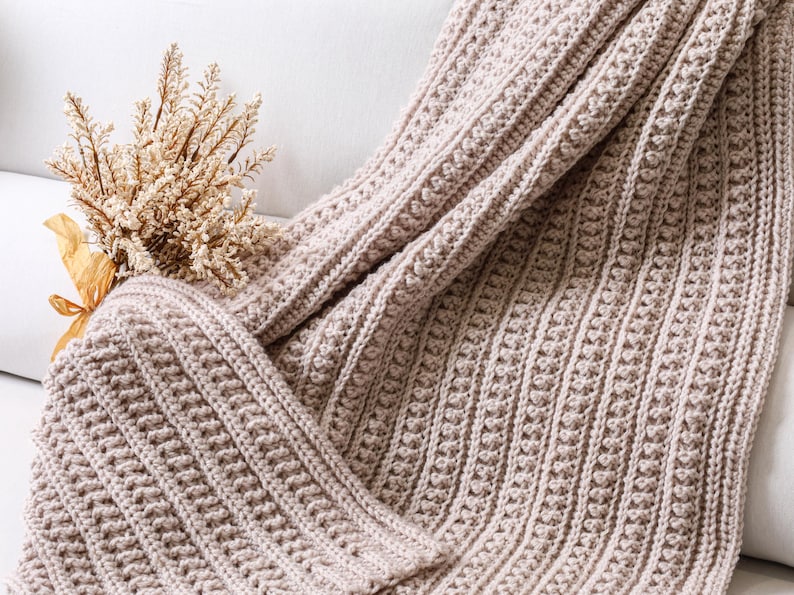 May include: A soft, beige crocheted throw blanket with a textured stitch pattern. The blanket is draped over a white couch.
