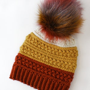 Puede incluir: Un gorro de punto con un pomp&oacute;n multicolor. El gorro est&aacute; hecho en tres colores: amarillo mostaza, naranja &oacute;xido y rojo burdeos.