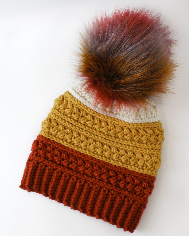 Puede incluir: Un gorro de punto con un pomp&oacute;n multicolor. El gorro est&aacute; hecho en tres colores: amarillo mostaza, naranja &oacute;xido y rojo burdeos.