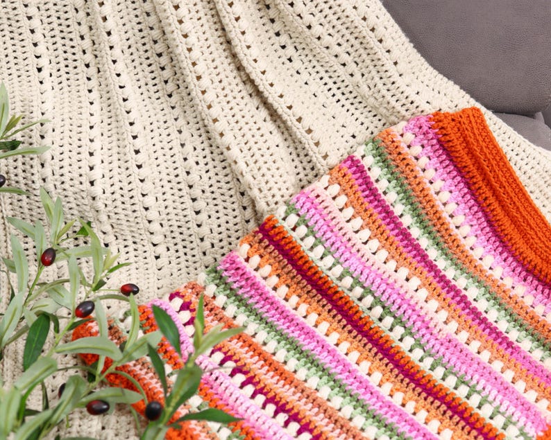 Pu&ograve; includere: Primo piano di due coperte all'uncinetto fatte a mano. Una &egrave; color crema con un motivo strutturato. L'altra &egrave; una coperta colorata con strisce rosa, arancioni, verdi e viola. Le coperte sono piegate e drappeggiate su una superficie.