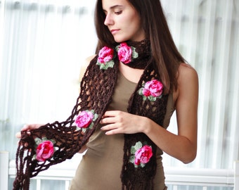 Può includere: Una sciarpa di uncinetto marrone con accenti di fiori rosa e verdi. La sciarpa è indossata da una persona con i capelli lunghi e castani.