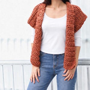 May include: A rust-colored, sleeveless crochet cardigan featuring a chunky, textured stitch pattern. The garment is open-front, with wide, ribbed-style lapels that drape down the torso, providing a layered look over a white shirt and blue jeans.