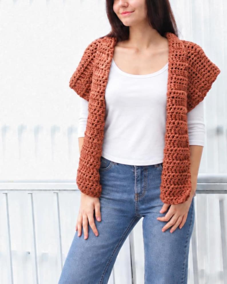 May include: A rust-colored, sleeveless crochet cardigan featuring a chunky, textured stitch pattern. The garment is open-front, with wide, ribbed-style lapels that drape down the torso, providing a layered look over a white shirt and blue jeans.
