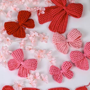 May include: A collection of crocheted bows in various shades of red and pink, including a large dark red bow, a medium light pink bow, and several smaller pink and red bows, arranged alongside small artificial light pink cherry blossom flowers.
