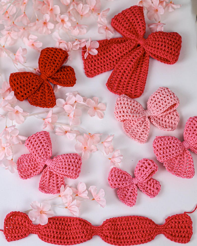 May include: A collection of crocheted bows in various shades of red and pink, including a large dark red bow, a medium light pink bow, and several smaller pink and red bows, arranged alongside small artificial light pink cherry blossom flowers.