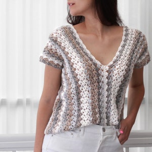 May include: A crocheted top with a v-neck and short sleeves. The top is made with a white, brown, and gray yarn and has a striped pattern.