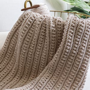 May include: A beige crocheted throw blanket with a textured pattern. The blanket is folded and draped over a white couch.
