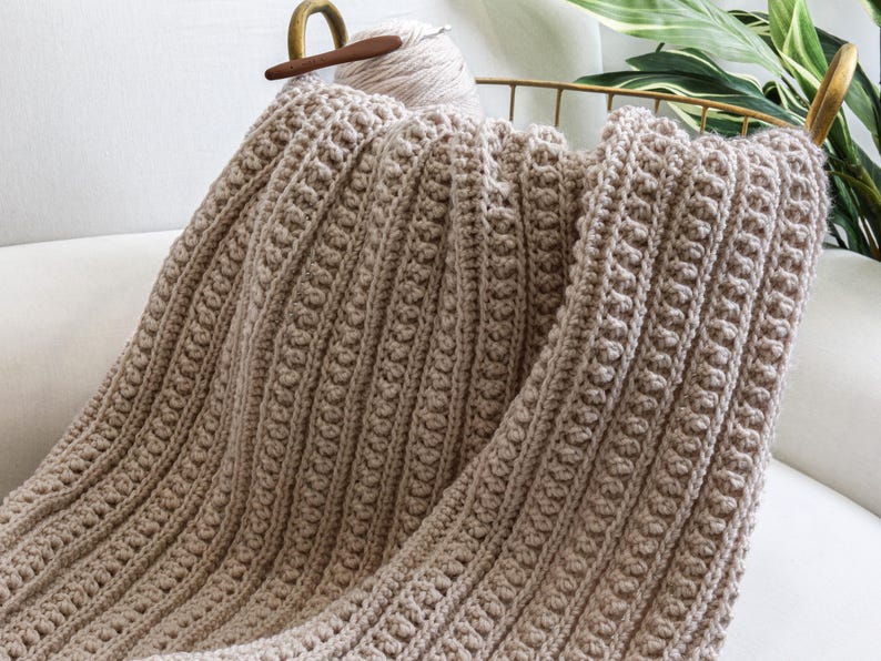 May include: A beige crocheted throw blanket with a textured pattern. The blanket is folded and draped over a white couch.