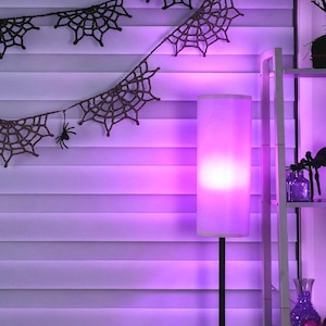 May include: A black crocheted spider web garland with small black spider figures hanging from the strands. A tall floor lamp with a cylindrical shade emits a bright purple glow, illuminating the room and the white horizontal window blinds.