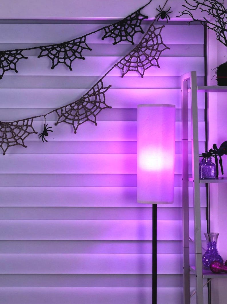May include: A black crocheted spider web garland with small black spider figures hanging from the strands. A tall floor lamp with a cylindrical shade emits a bright purple glow, illuminating the room and the white horizontal window blinds.