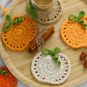 May include: A collection of round, crocheted pumpkin-shaped coasters in orange, light orange, and cream yarn, each featuring a green crocheted stem and leaf. The set includes a bundle of cinnamon sticks and two small brown acorns as accents.