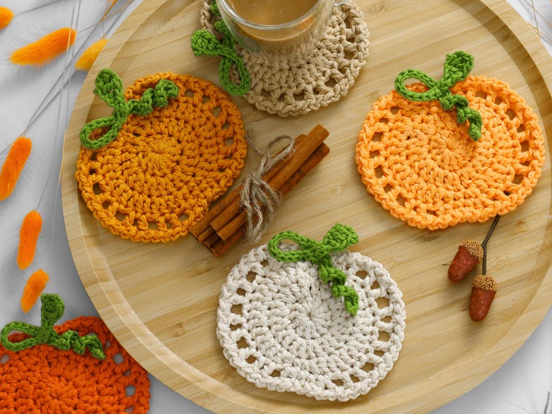 May include: A collection of round, crocheted pumpkin-shaped coasters in orange, light orange, and cream yarn, each featuring a green crocheted stem and leaf. The set includes a bundle of cinnamon sticks and two small brown acorns as accents.
