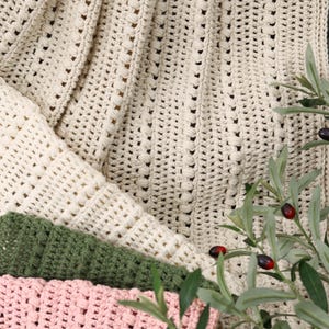 Pu&ograve; includere: Primo piano di diverse coperte all'uncinetto in vari colori. Le coperte sono color crema, verde e rosa. La coperta color crema &egrave; drappeggiata sopra le altre, con un motivo strutturato. Rami di ulivo con foglie verdi e olive nere in primo piano.