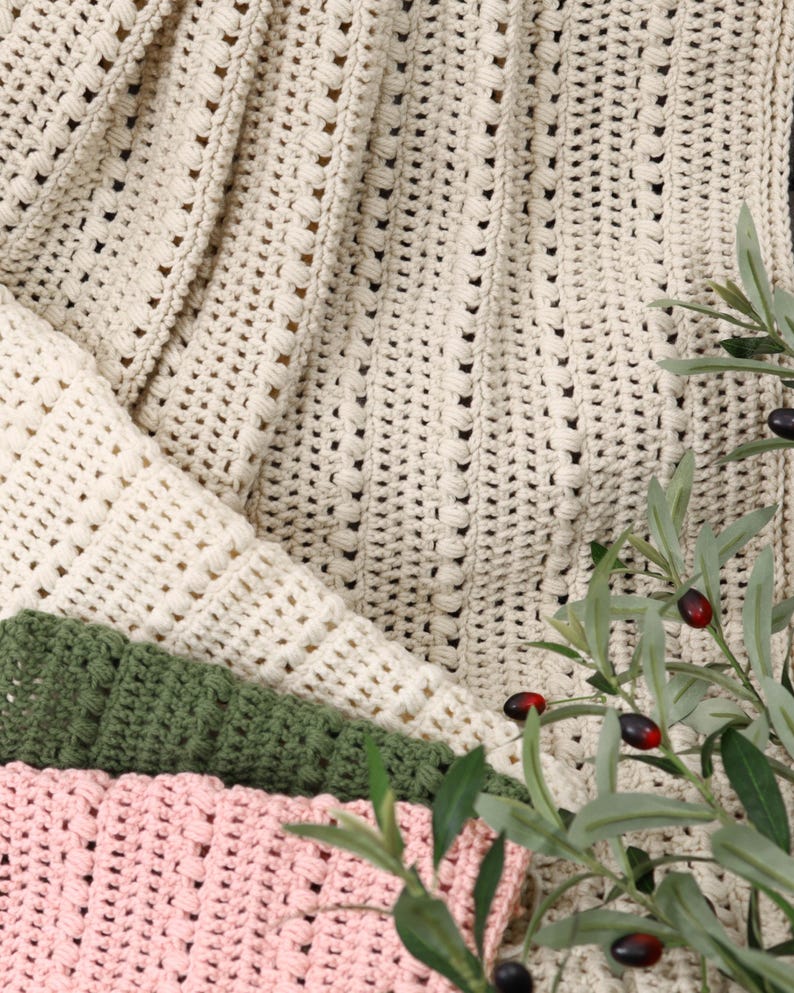 Pu&ograve; includere: Primo piano di diverse coperte all'uncinetto in vari colori. Le coperte sono color crema, verde e rosa. La coperta color crema &egrave; drappeggiata sopra le altre, con un motivo strutturato. Rami di ulivo con foglie verdi e olive nere in primo piano.
