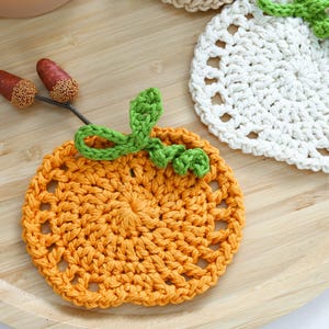 May include: A round orange crochet coaster with a green yarn stem and leaf detail, accompanied by two small brown acorns on a thin twig. A second white crochet coaster with a green stem is partially visible in the upper right corner.