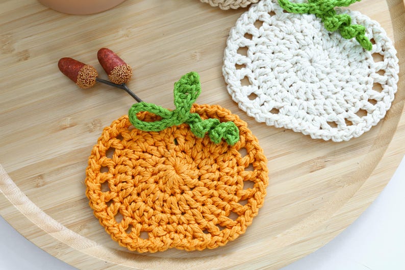 May include: A round orange crochet coaster with a green yarn stem and leaf detail, accompanied by two small brown acorns on a thin twig. A second white crochet coaster with a green stem is partially visible in the upper right corner.