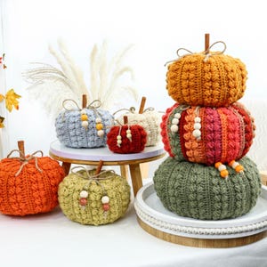 May include: A collection of handmade crochet pumpkins in various sizes and colors including orange, light blue, red, cream, olive green, and a multi-colored stacked pumpkin tower. Each pumpkin features a wooden stem and decorative wooden beads.