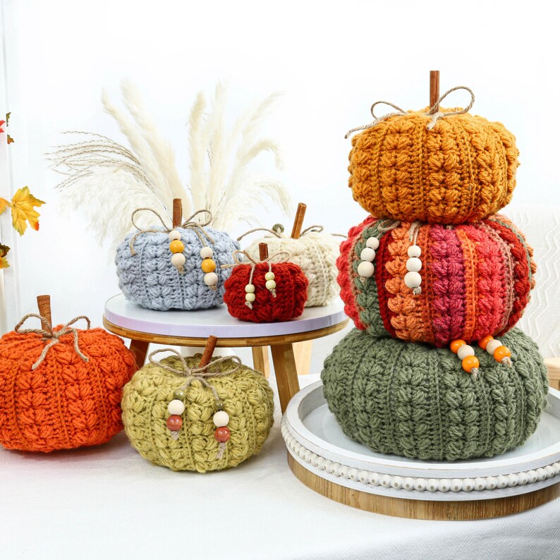May include: A collection of handmade crochet pumpkins in various sizes and colors including orange, light blue, red, cream, olive green, and a multi-colored stacked pumpkin tower. Each pumpkin features a wooden stem and decorative wooden beads.